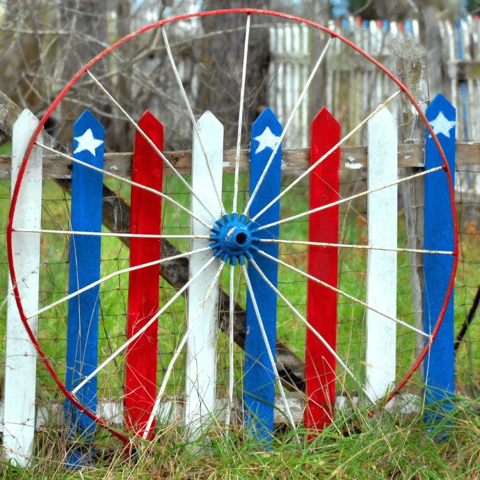 A rustic wooden fence is painted in red, white, and blue stripes with stars, resembling the American flag. An old wagon wheel leans against the fence, and grass grows in the foreground.