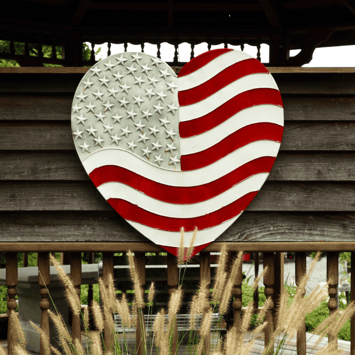 A heart-shaped decoration painted with the American flag hangs on a wooden wall, with ornamental grass in the foreground and a gazebo structure in the background.