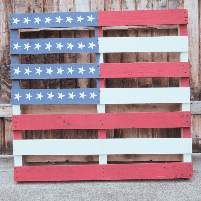 A wooden pallet painted to resemble the American flag, with red and white stripes and a blue section in the top left corner featuring white stars, leaning against a wooden wall.