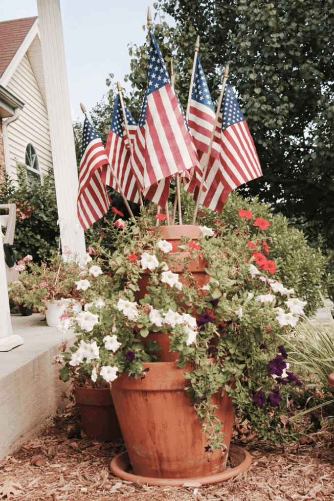 Several small American flags are displayed in flower pots filled with blooming petunias and other flowers on an outdoor porch, with a house and trees in the background.