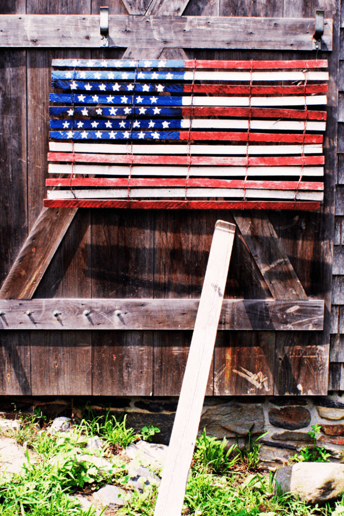 A rustic wooden American flag hangs on a weathered barn wall above a patch of grass and stones, with a wooden plank leaning against the structure.
