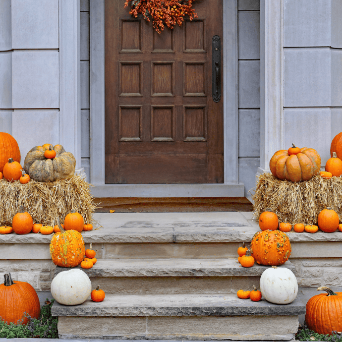 A front porch decorated for autumn with hay bales, orange and white pumpkins, gourds, and a fall wreath on a wooden door. The decorations are arranged on the steps and around the entrance.