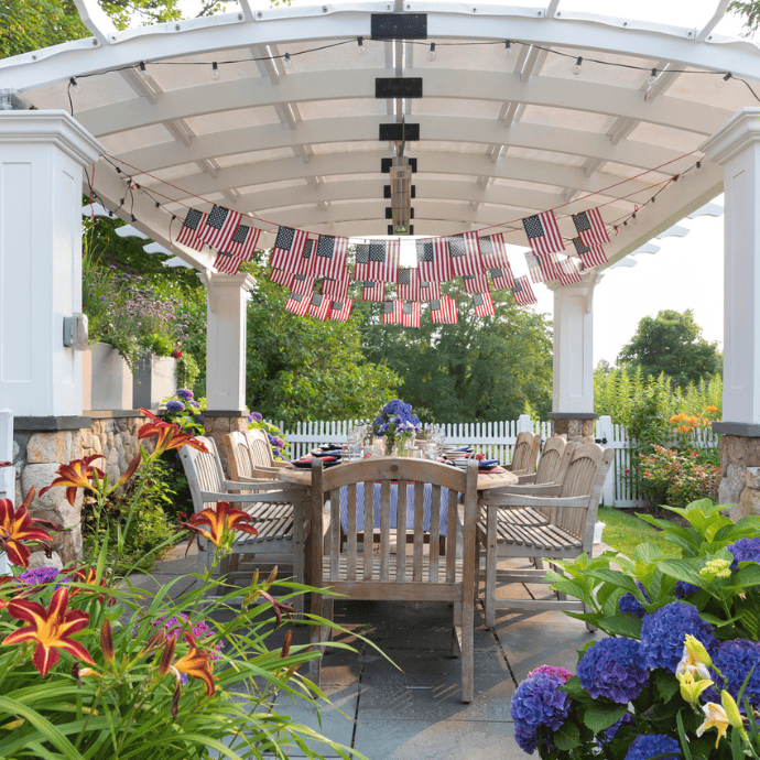 A wooden outdoor dining table sits under a white pergola adorned with easy 4th of July patriotic decorations, like hanging American flags, surrounded by colorful flowers and greenery in a sunny garden setting.