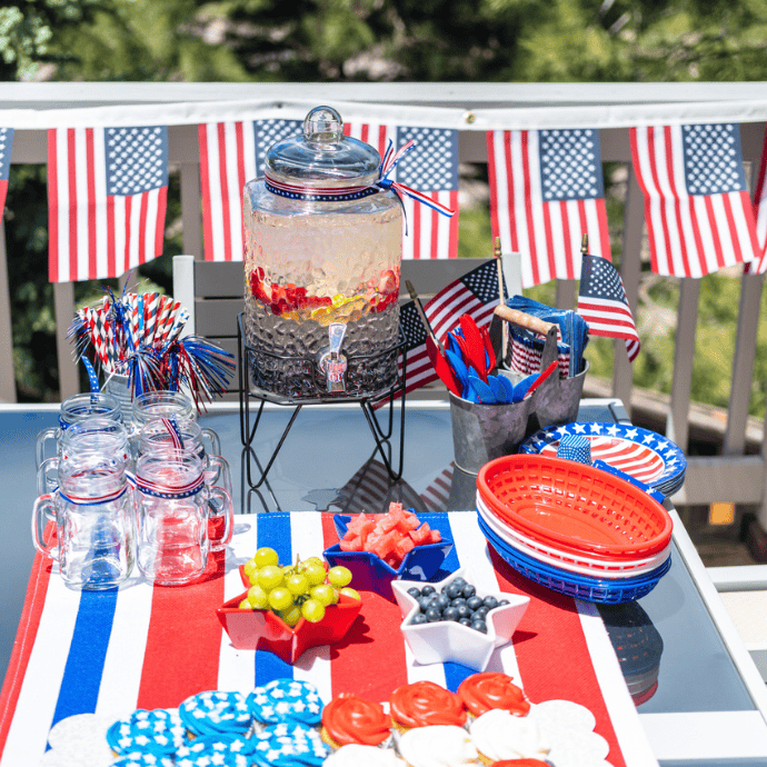 A festive outdoor table is set for a 4th of July celebration with patriotic decorations—American flags, patriotic plates, easy decorations, snacks, and a large drink dispenser. Red, white, and blue colors dominate the tableware and decor.