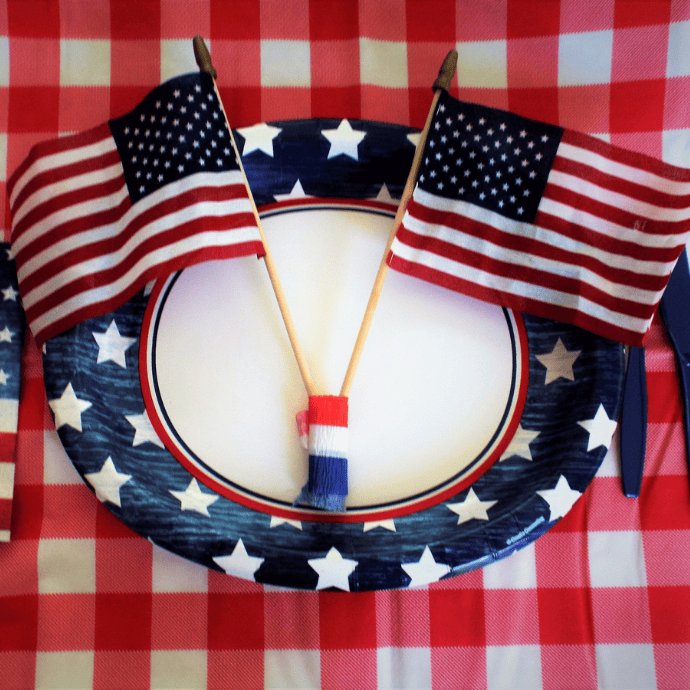 An oval paper plate with a blue and white star border holds two small American flags and a red, white, and blue decoration—easy patriotic decorations perfect for your 4th of July table, set on a red and white checkered tablecloth.