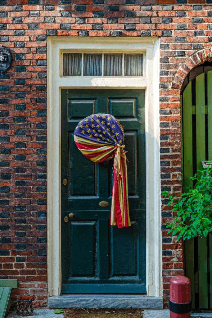 A dark green door decorated with a large fabric bow in the design of the American flag—perfect to celebrate the 4th of July—set in a brick wall with a small window above and some greenery to the right side.