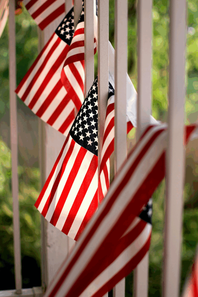 Small American flags, perfect as patriotic decorations, are displayed vertically along a white railing with a green, blurred background. The focus is on the closest flag, highlighting the stars and stripes—ready to celebrate the 4th of July.