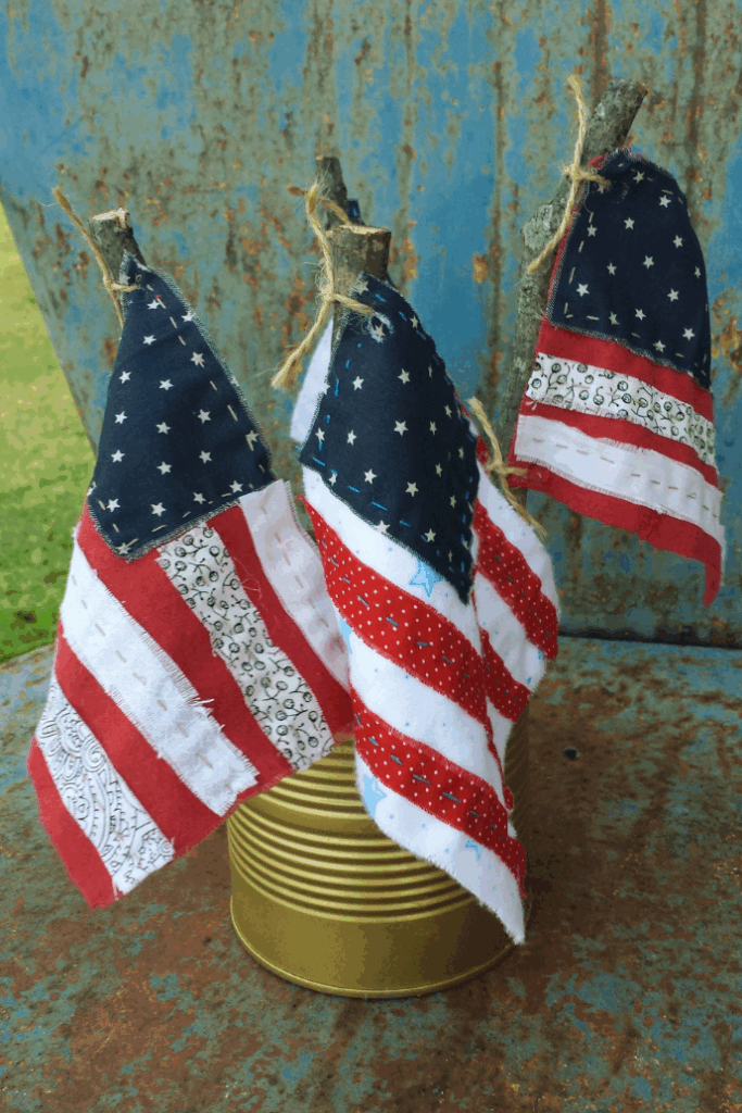 Three small homemade American flags made of fabric scraps are attached to sticks and displayed in a gold tin can, making easy decorations for 4th of July celebrations, set against a rustic, weathered blue metal background.