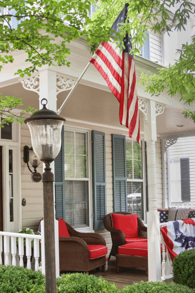 A cozy front porch with wicker chairs featuring red cushions, easy patriotic decorations for the 4th of July, an American flag hanging above, blue shutters on the windows, and a vintage-style lamp post amid lush green bushes and trees.